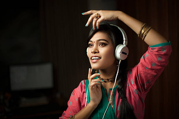 Traditional Hindu teenage girl listening music through headphone and dancing. Indoor low key image of a fashionable Asian Hindu teenage girl listening music through headphones and dancing at night in her domestic room. She is in traditional Hindu dress that is Salwar Kameez and dupatta. Horizontal composition with copy space and selective focus. indian music stock pictures, royalty-free photos & images