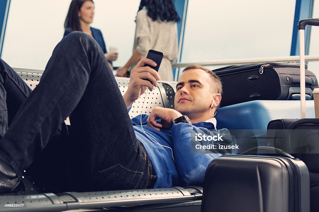 Waiting for the flight Focus on the man lying down on bench at the airport lounge, wearing earphones and using smart phone. Airport Departure Area Stock Photo