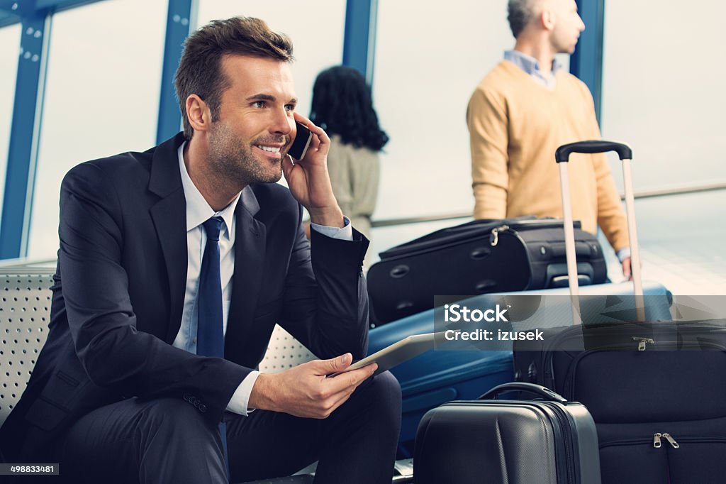 Man on phone at the airport Smiling businessman talking on his mobile phone in an airport lounge.  The man is sitting on the edge of his seat with his phone in his left hand and a digital tablet in his right hand.  He is wearing a black jacket, black pants, a white shirt and a black tie.  There are four suitcases to the left of the image; two of the suitcases are standing upright and two are lying down.  There are other people in the background looking out of the window, waiting for their flight to arrive. Airport Stock Photo