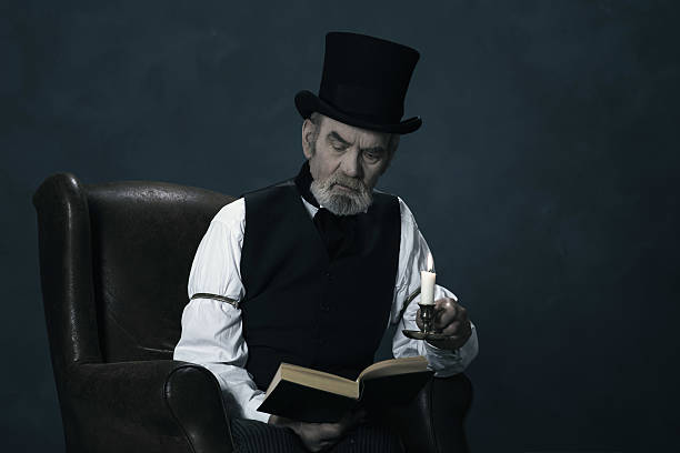 dickens scrooge homme assis dans un fauteuil de lecture de livre à la lueur des bougies. - scrooge photos et images de collection