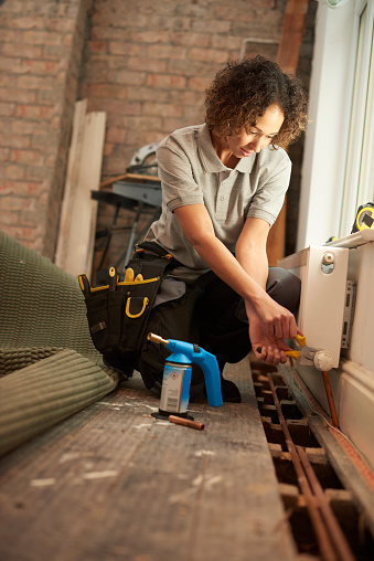 a mid adult female plumber is installing a new radiator into a property undergoing refurbishment. The floorboards are raised with new paperwork shown , and she is tightening the valve on the new radiator. She is wearing work trousers , and tool belt . In the background more work tools can be seen as is an exposed brick wall .