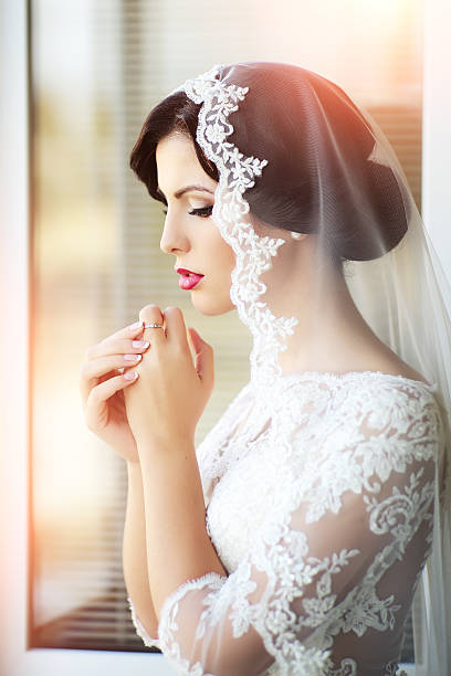 Pensive brunette bride Portrait with side view of one beautiful young brunette sensual pensive bride in white lace dress and veil on head holding hands near face, vertical picture wedding ceremony formalwear people clothing stock pictures, royalty-free photos & images