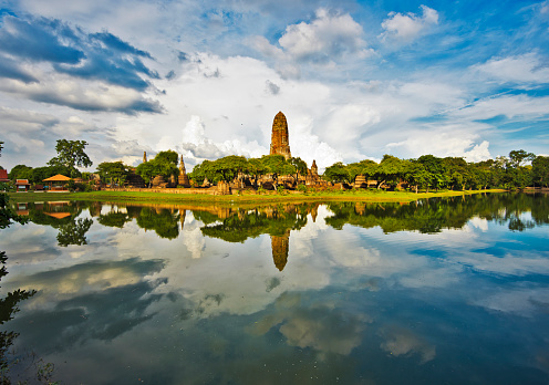 Southwest panoramic view of Wat Phra Ram temple complex in Ayutthaya historical park in Thailand, with the Prang tower in the middle, the before thunderstorm cloudscape is at background and the lake mirror surface at foreground