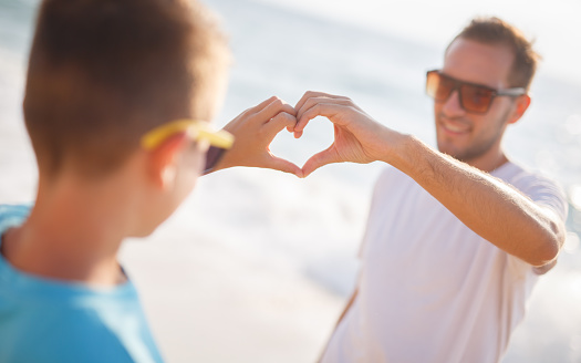 Father and son enjoy on the beach. Heart symbol made with hands.
