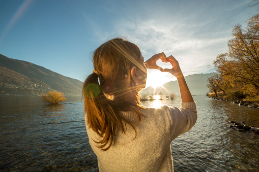 Young woman by the lake making a heart shape finger frame. Lake and mountain landscape. Swiss Alps on the background.