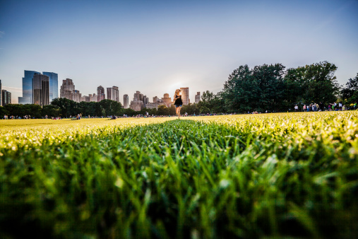 Teenage girl runs in front of Manhattan skyline at the Sheep Meadow, Central Park, New York City. People relaxing ath the grass at evening. 