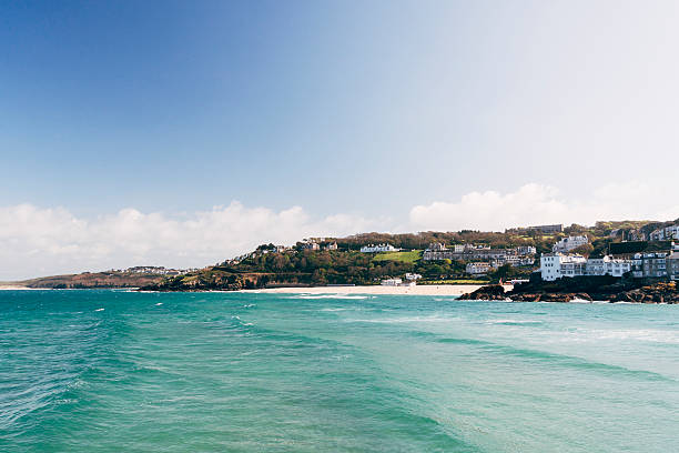 St Ives coastline from the harbour wall in Cornwall, England. Porthminster beach is towards the right edge of the frame and Carbis Bay toward the left edge of the frame. In the foreground waves are rolling across the blue sea.