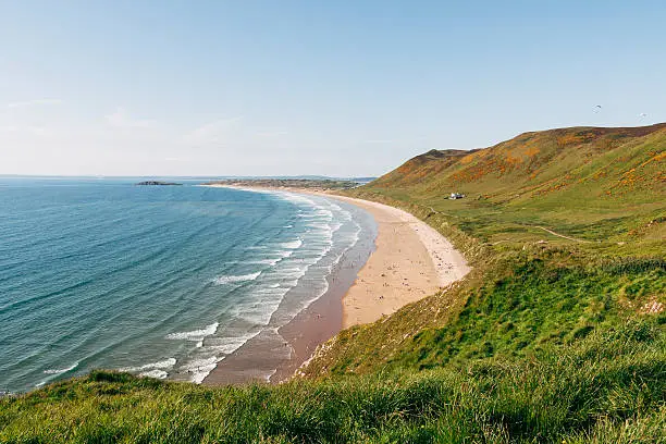 Rhossili Bay and beach on the Gower peninsula on the coast of South Wales. It is close to Swansea and is designated as an area of outstanding natural beauty in the United Kingdom. Large hills roll down to the sandy beach and sea on a beautiful sunny day.