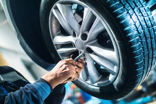 Closeup of unrecognizable male mechanic unscrewing the bolts on the rim of car wheel in order to replace tyre or the wheel itself. After he got those bolt loose with power tool he's using hand screwdriver to pull them out. The mechanic is wearing blue working uniform and the wheel is lit with blue fill light. The car is lifted up on a car jack