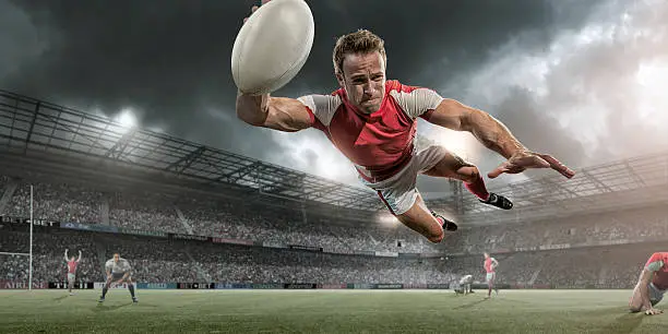 A low angle view of professional rugby player diving in mid air with rugby ball to score a try. The action takes place in on a outdoor rugby pitch in a generic stadium full of spectators under a dark stormy sky at sunset. All players are wearing generic rugby kit.