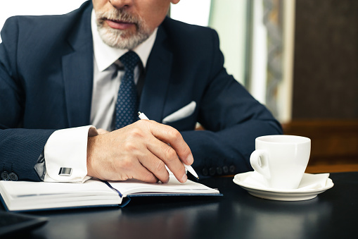 Close up photo of businessman in expensive and classic office. Aged businessman wearing suit, making notes and having coffee