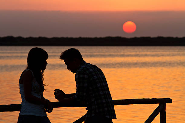 A girl and a guy are talking in the sunset. Their silhouette comes off on a wonderful sunset ober the lake