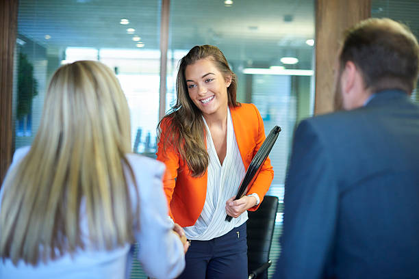 job interview first impressions a young graduate walks into an interview room full of confidence and positivity energy . She is holding her cv and smiling at the interview panel before her. She is wearing blue trousers with an orange suit jacket , as she opens the door and strides in and shakes hands with a woman on the panel . in the foreground we can see the back of two of the panel , a man and a woman. Trainee stock pictures, royalty-free photos & images