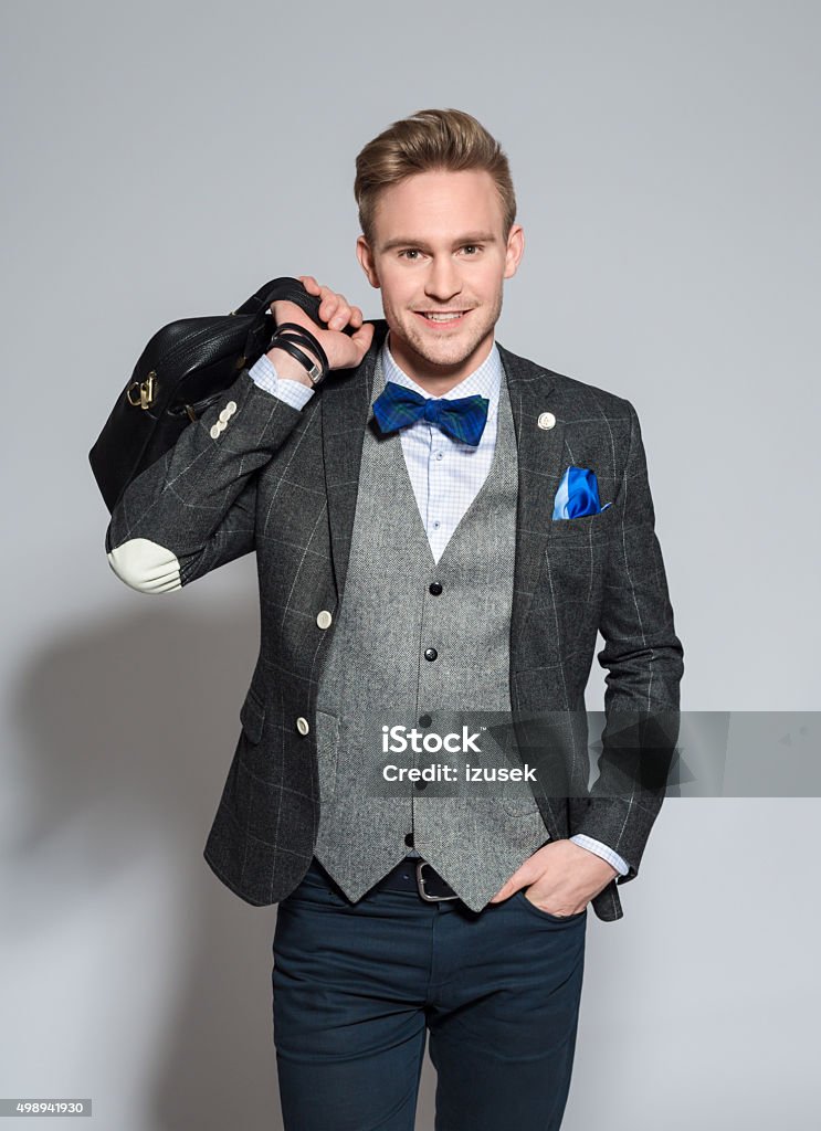 Charming young businessman wearing tweed jacket and bow tie Portrait of fashionable, elegant young businessman wearing tweed jacket and bow tie, holding leather briefcase, smiling at camera. Studio shot, one person, grey background. 2015 Stock Photo