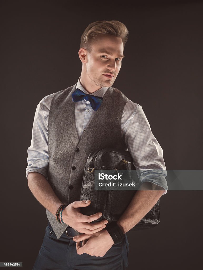 Handsome elegant man wearing bow tie, holding bag Fashionable elegant man wearing tweed vest, shirt and bow tie, holding a bag, looking at camera. Dark tone, black background. Briefcase Stock Photo