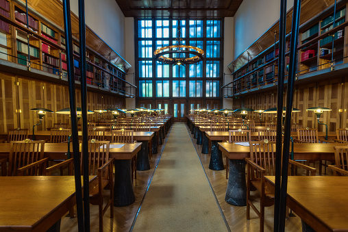 Rows of antique wooden tables and chairs in a public library reading room with shelves, windows and a chandelier in the background.