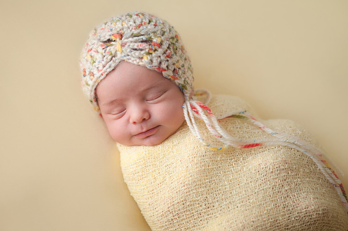 A portrait of a beautiful, two week old, newborn baby girl wearing a crocheted bonnet. She is smiling and sleeping on yellow colored fabric.