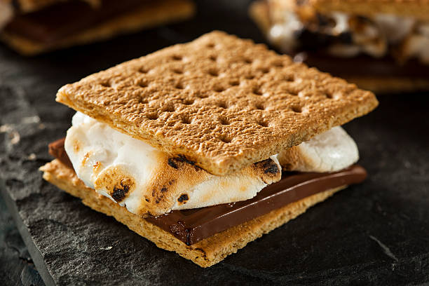 S'more with burnt marshmallow and melted chocolate Close up image of S'mores dessert with roasted marshmallows and a chocolate bar sandwiched between two square graham crackers.  The dessert is displayed on a dark gray background, with two more S'mores in the background.  The background is slightly out of focus. smore photos stock pictures, royalty-free photos & images
