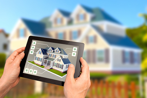 Choosing a new house to buy: hands holding a tablet showing a real estate listing with model of a new country home to buy, with labels pointing at features, and the potential house deal on the background. Buying dream house, real estate business and investment, home automation.