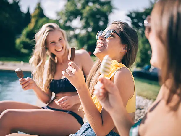 Teenaged girl friends sitting next to a pool on a sunny summer day laughing and eating ice creams
