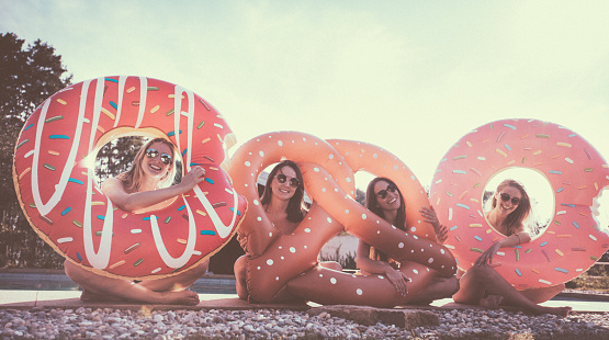 Portrait of four girl friends sitting next to a pool and holding funny pool inflatables made to look like donuts and a pretzel, smiling happily at the camera outdoors on a summer day