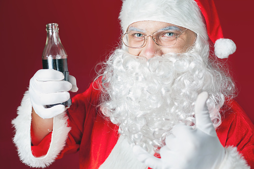 Funny Santa Claus holding Coca Cola fresh beverage at glass bottle. Christmas holiday, New Year. Thumb up gesture. Copy space for disign, text at red background