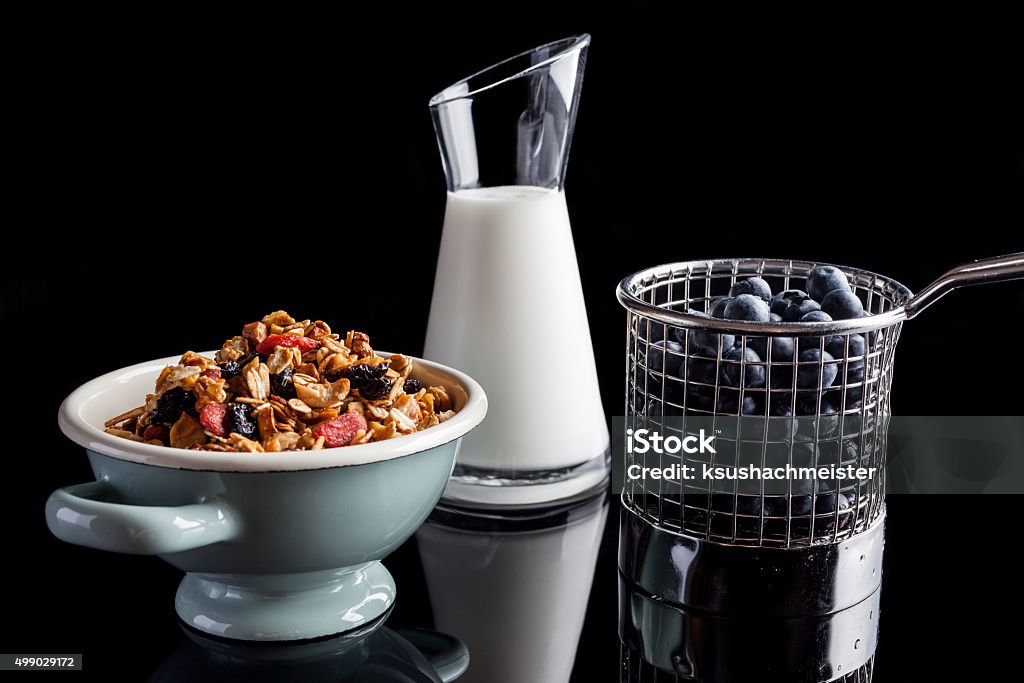 Blueberries granola and yogurt on black from side Blueberries in steel cup stainer, granola in an enamel white and blue bowl and yogurt in a glass jug on black background from side 2015 Stock Photo