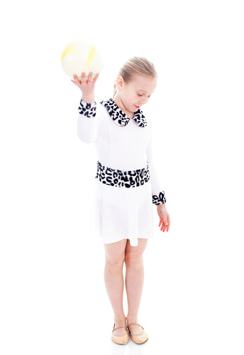Little girl gymnist with a ball, standing and looking down. Wearing a white dress with lepard  belt.