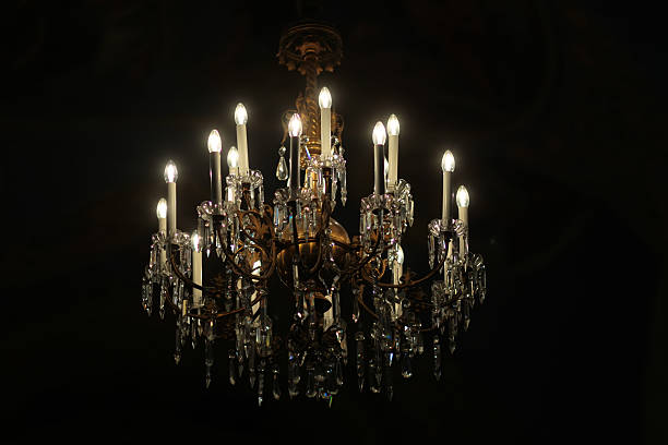Photo closeup one classic crystal cut-glass grand chandelier lighting fixture ceiling lamp with electric opal bulbs pendants switched on illuminating darkness over black background, horizontal picture
