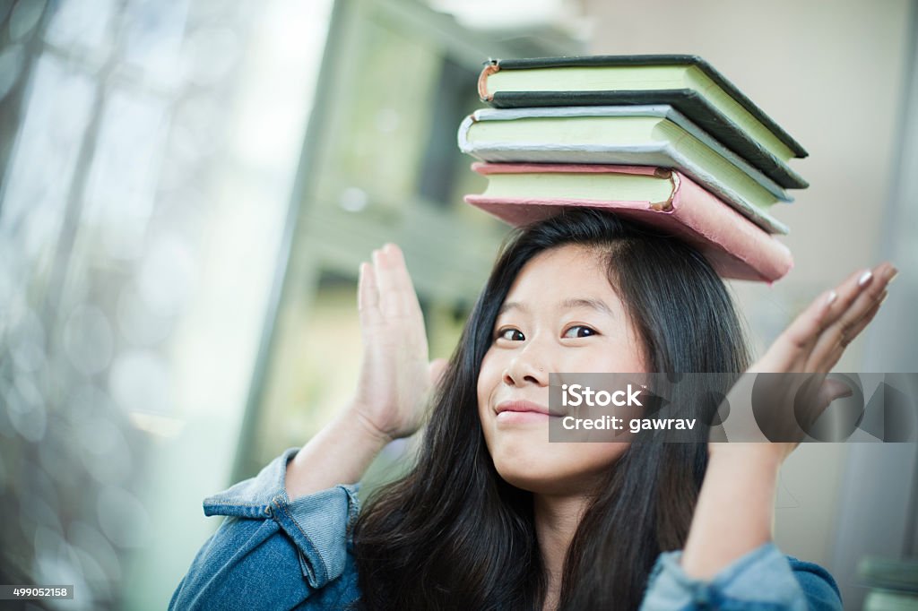 Happy Asian girl student balancing stack of books on head. Indoor head and shoulder image of a happy Asian teenage girl student with long hair wearing denim shirt balancing the stack of books on her head and looking at camera with her hands up. She is confident for her upcoming university or high school exams as she has prepared herself by studying hard. Horizontal composition with copy space and selective focus. Book Stock Photo