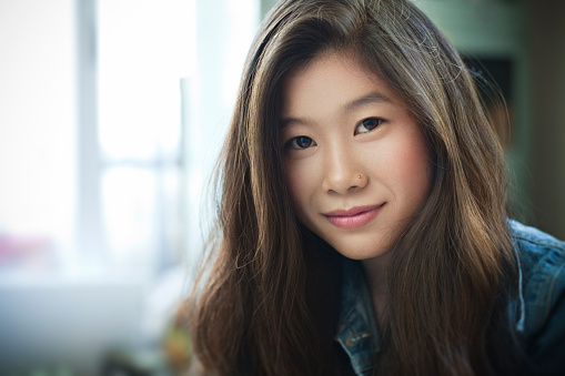 Day time indoor image of beautiful, happy late teen girl or young woman with long brown hair of Asian ethnicity looking at camera. Hade and shoulders, horizontal image with copy space and selective focus.