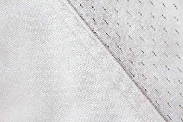 Full frame image of clean, white, jersey material.  There is a seam between the solid fabric, and the breathable fabric with holes in it.  The seam is diagonal.