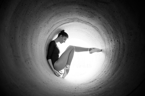 Young ballerina dancing and posing in tunel with strong  light at it end. Woman is very attractive and flexible and she wearing dark leotard and ballet slippers. 
