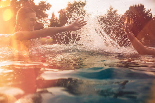 Summer swimming pool with girls splashing water playfully Backyard swimming pool on a late summer evening with girls splashing each other with water while play fighting, with colourful sun flare vintage pool stock pictures, royalty-free photos & images