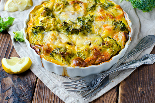 A colorful fish and broccoli casserole in a white baking dish.  The casserole sits on a plank style wooden surface with lemon wedges and forks.