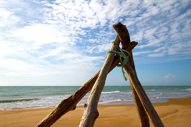 Driftwood teepee tied with rope on a golden beach in the foreground with the Mediterranean Sea and blue puffy-cloud sky in the background. Shot in Sicily. Copy space available.