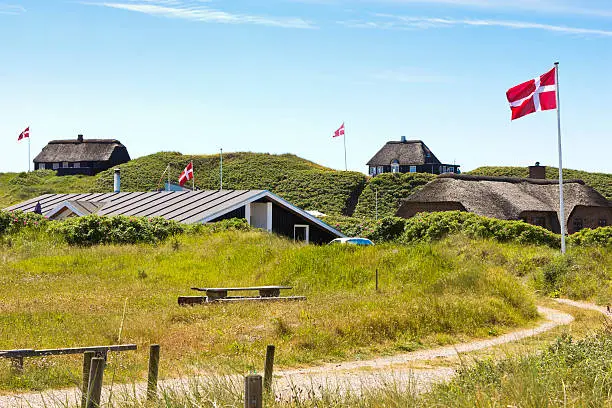 Summerhouses at the western Jutland, Denmark. Specifically for Denmark, the danish flag "Dannebrog" is used intensively by many private houses. The houses in the image are located close to the western sea "Vesterhavet", in Jutland, which is a prime area of vacations for the danes as well as for a large part of the tourists. The flags are waving in a light summer breeze under a blue sky with a few scattered clouds. The houses are thatched as is customary for the area.