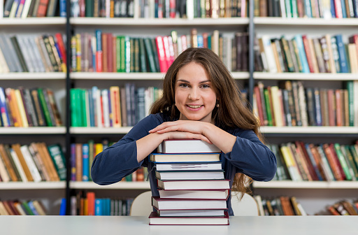 smiling young girl sitting at a desk in the library with her arms on a big pile of books and chin on them, looking in front of her, thinking,  a concept of knowledge, blurred books at the background