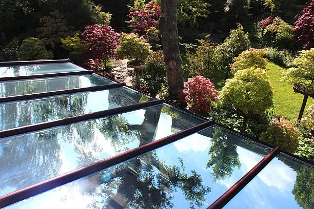 Photo showing a large glass conservatory roof with brown UPVC dividers, with the individual panels  panes of glass showing reflecting the blue sky and the trees in the surrounding garden.