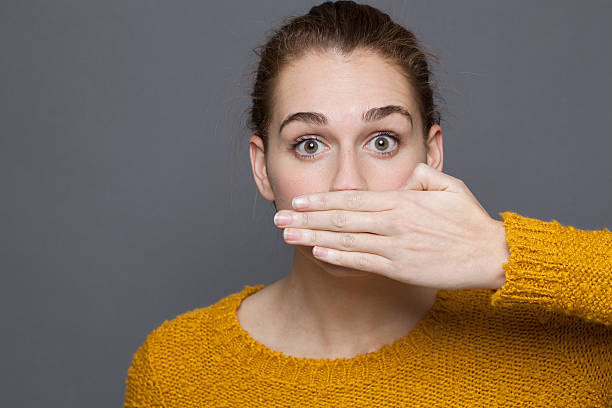 stunned young woman covering her mouth for silence negative feelings concept - portrait of amazed beautiful 20s girl covering her mouth for gag metaphor,studio shot on gray background concepts topics stock pictures, royalty-free photos & images