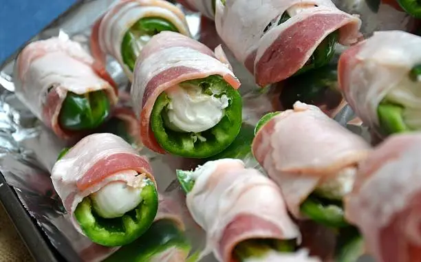 A dslr color macro image of fresh cut and deseeded jalapeno peppers, stuffed with cream cheese then wrapped with bacon. Raw food state, placed on a foil lined baking sheet ready for baking in the oven. Natural lighting, studio shot.