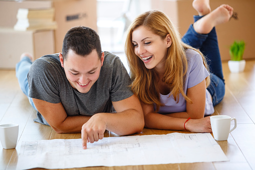 A young heterosexual couple is lying down on the floor with an architectural drawing of their new apartment in front of them. The couple is smiling and looking at blueprint while the man is showing something on the drawing. They are making plans how to decorate their house and where to put the new furniture. In the background, in blur, we can see the home interior.