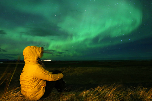 A woman wearing a bright yellow winter jacket is sitting in a field as she is watching a Northern Lights display during a strong aurora storm in Southern Iceland in November 2015. Canon EOS 5Ds, f/4, 6sec, ISO-2500.