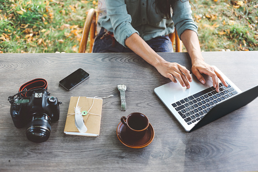 female novelist, blogger, freelancer or a photographer, typing on the laptop keyboard while having a cup of coffee, sitting in the bright outdoors at the desk. Retro processed with vibrant colors. Taken with a large aperture prime lens for shallow depth of field.