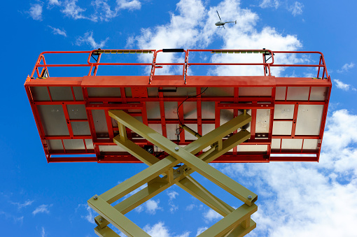 Scissor lift platform with hydraulic system at maximum height range painted in orange and beige colors, large construction machine, heavy industry, white clouds and blue sky on background 