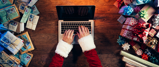 Santa working at desk and typing on a laptop surrounded by colorful Christmas gifts and letters, hands top view