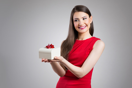 A photo of young woman handing white gift box with red bow. She's smiling. She's wearing red, elegant dress.