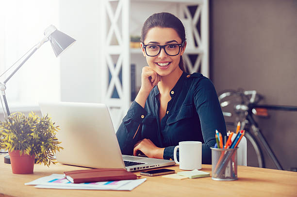 Smart and confident. Attractive young woman in eyeglasses holding hand on chin and smiling while sitting at her working place in office secretary stock pictures, royalty-free photos & images