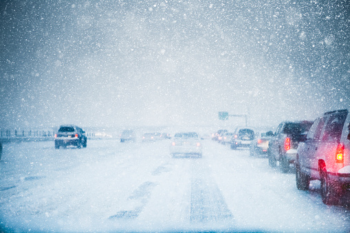Winter weather and slow moving traffic creeping along a snow covered and icy freeway in a winter snow storm blizzard. Taillights are on in low light and low visibility road conditions. Copy space available. High resolution color photograph with horizontal composition.