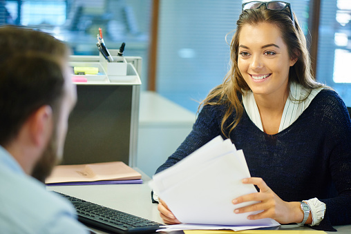 A young female office worker chats to her supervisor across a desk in a busy office .